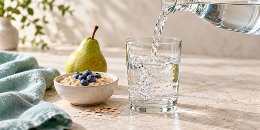  Water being poured into a clear glass beside a bowl of cooked oats topped with blueberries and a pear.
