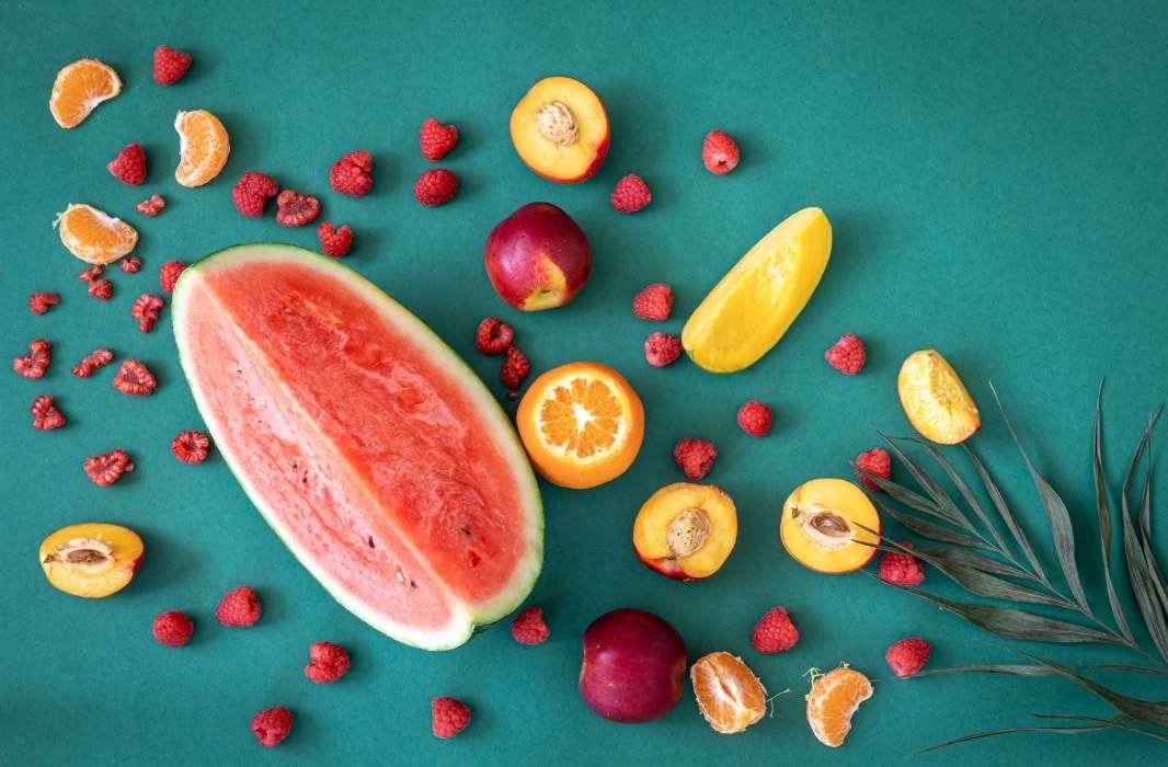 Flat-lay of colorful fresh fruits including watermelon, citrus, and berries on a teal background, representing natural antioxidants and skin-nourishing nutrients.