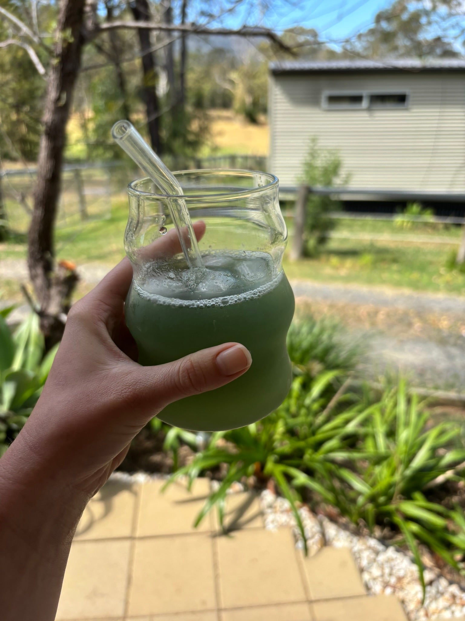Hand holding a glass jar with a green liquid and straw outdoors.