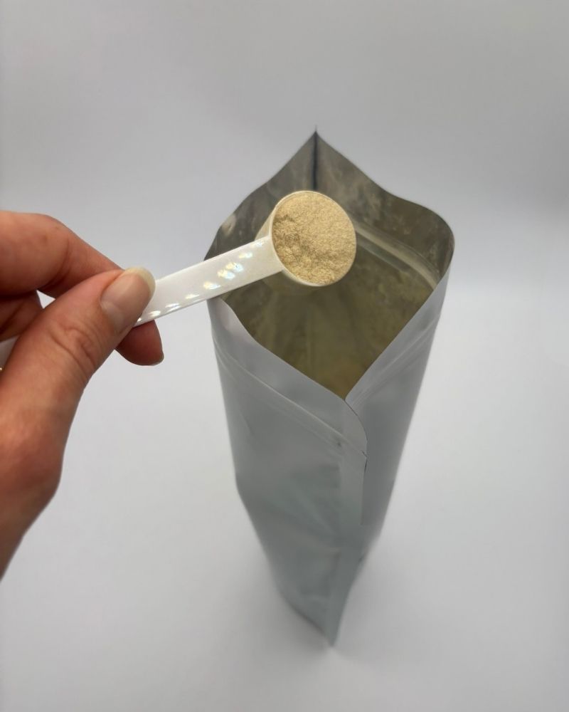 Hand holding a spoonful of powder above a gray bag on a light gray background