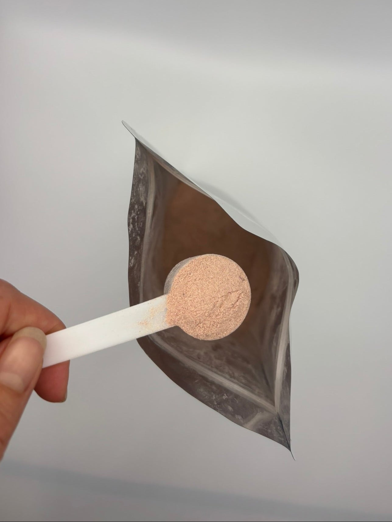 Hand holding a spoon with a brown powder over a clear bag on a light gray background