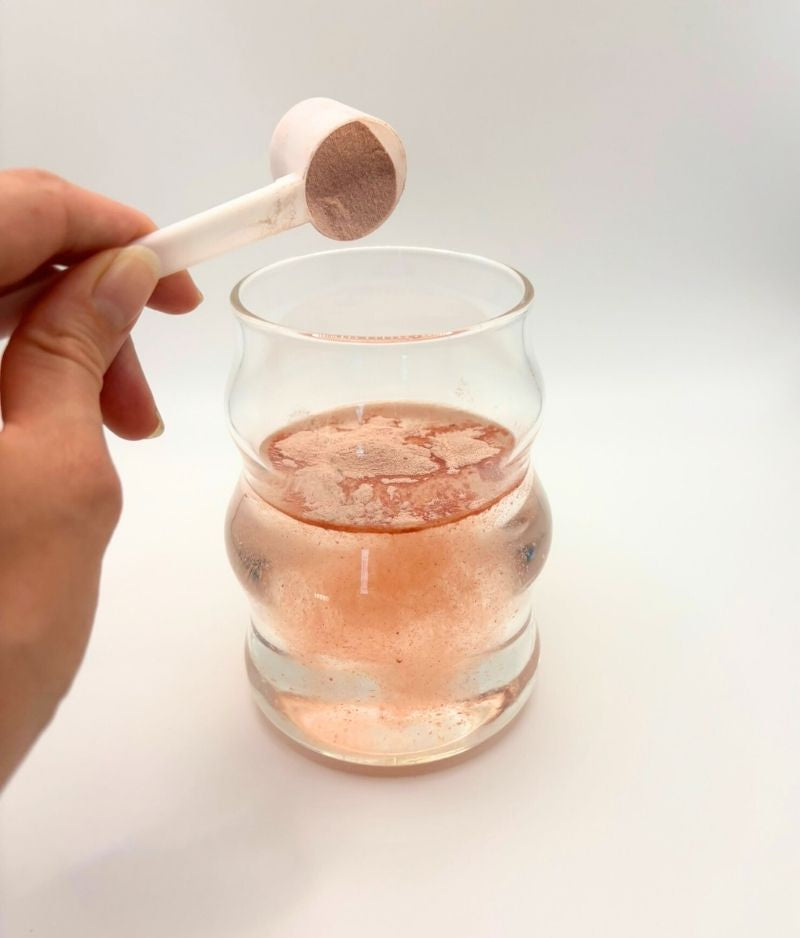 Hand holding a plastic scoop of custom skin glow powder over a glass of pink liquid on a white background